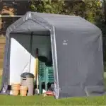 Stranski pogled na siv skladiščni šotor Maxy velikosti 3m x 3m delno odprt ki razkriva zložene stole in vrtno opremo obdan z bujnim zelenjem