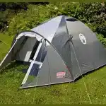 Šotor za kampiranje Coleman Crestline 3 postavljen na zeleni travi s sivo in belo barvno shemo ter vidnim logotipom blagovne znamke kar kaže na robustno zasnovo in kakovostno znamko primerno za kampiranje v naravi