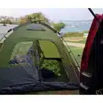 Šotor za kampiranje Instant Dome 5 postavljen ob morju z odprtim vhodom ob strani rdečega vozila poudarja enostavnost postavitve in uživanje v naravi