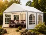 White outdoor party tent with arched window panels and a wooden sectional sofa with burgundy and patterned cushions in a sunlit garden setting