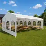 White marquee tent on a grassy lawn with arched window panels and long wooden tables and benches inside under a blue sky and trees