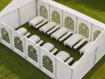 A white event tent on green grass with long rectangular tables and benches arranged in rows inside arch windowed walls as seen from above