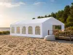 White tent on a sandy beach beside a wooden deck with outdoor seating ocean and trees in the background