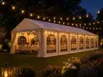 White marquee tent illuminated by string lights at night with round tables and chairs set for an outdoor event on a lawn