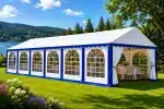Large white event tent with blue trim set up on a grassy lawn by a lake and forested hills tables and chairs inside